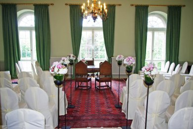 Schloss Stülpe großer Saal mit grünen Vorhängen, einem Kronleuchter, Stühlen mit weißen Hussen, Blumen und einem großen Tisch vor einem Fenster mit zwei antiken Stühlen davor